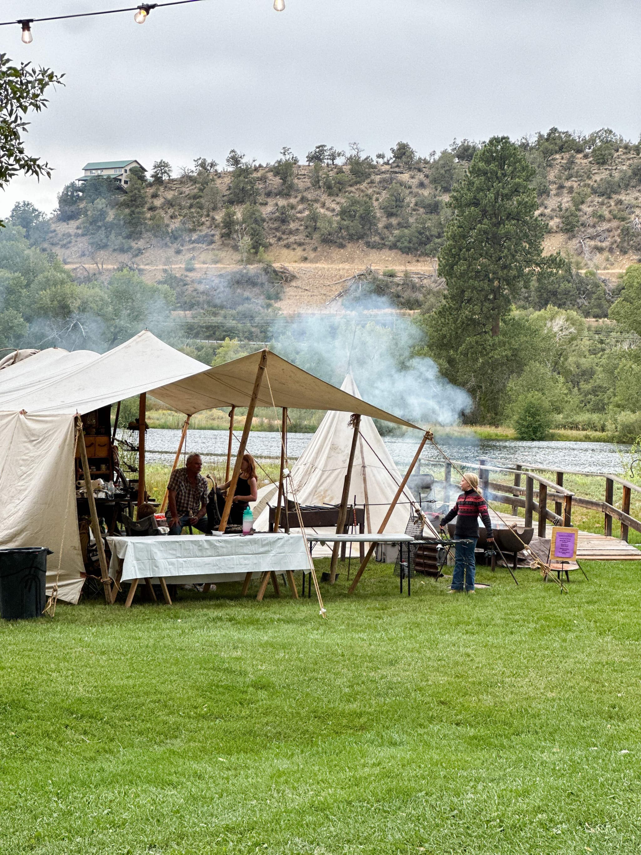 Chuckwagon camp scene with cooking tents and smoke rising by the pond