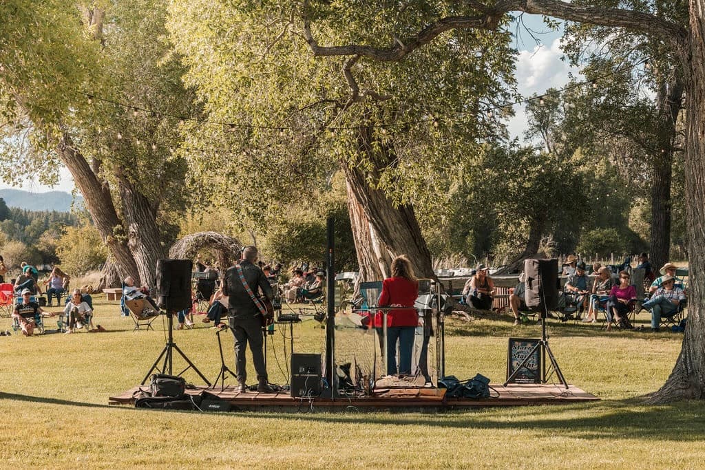 Musicians performing on the outdoor stage beneath towering cottonwood trees as the crowd relaxes on the lawn at Locals Night