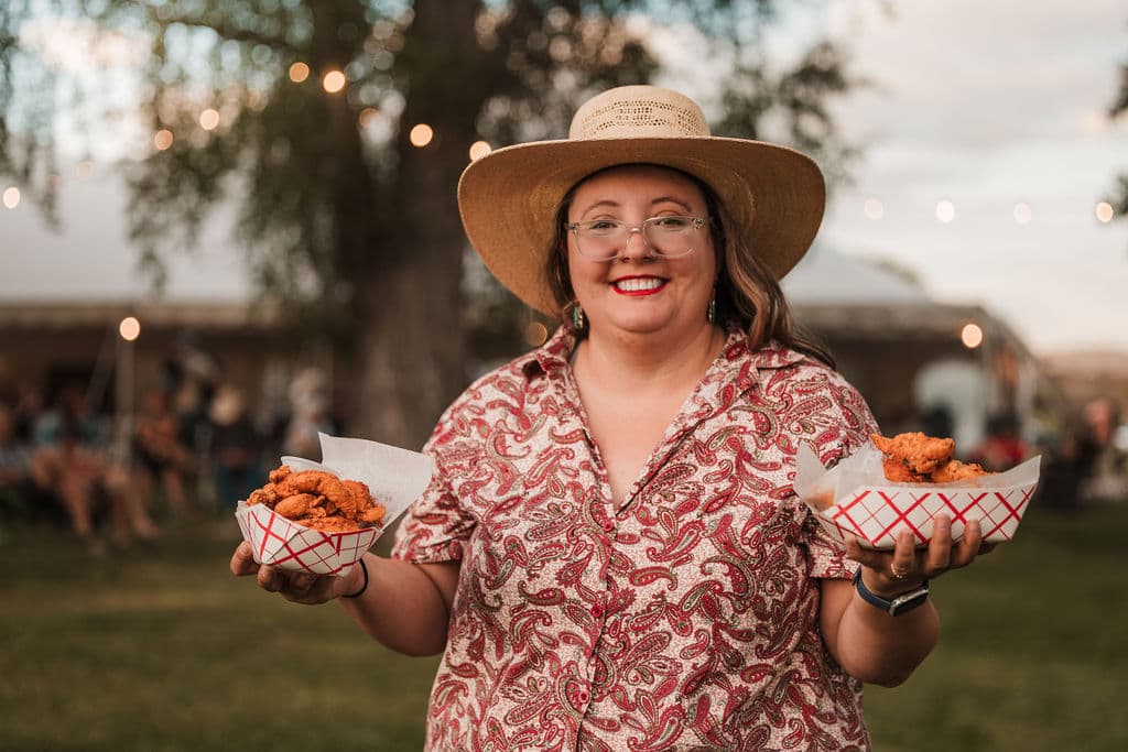 A guest smiling while holding two baskets of fresh food beneath the string lights