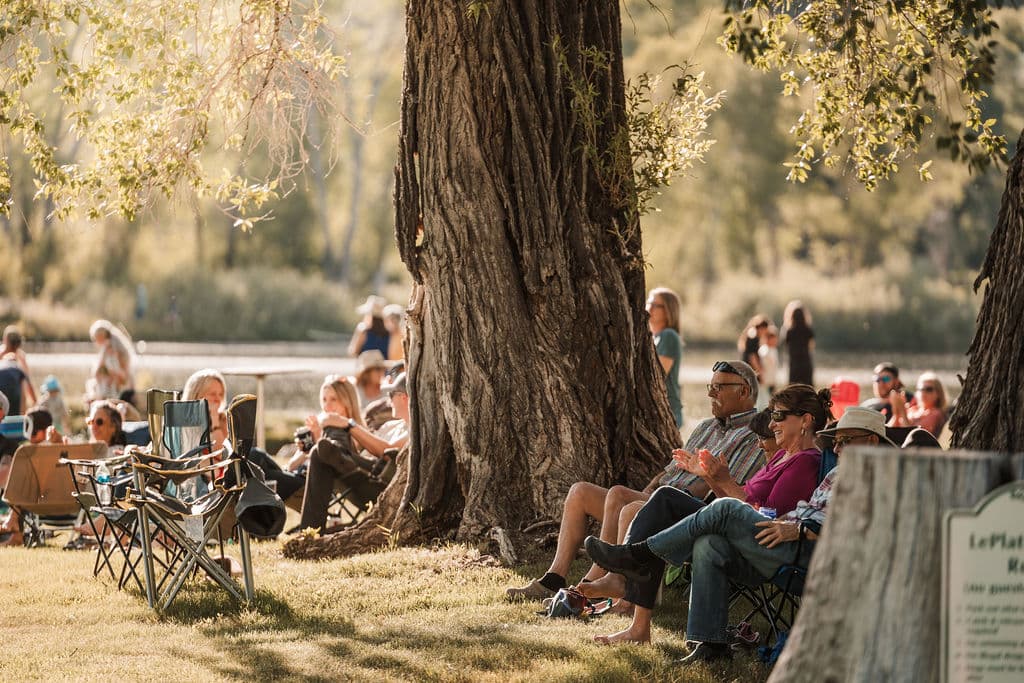 Guests gathered in lawn chairs beneath a grand cottonwood tree, clapping and enjoying live music by the pond