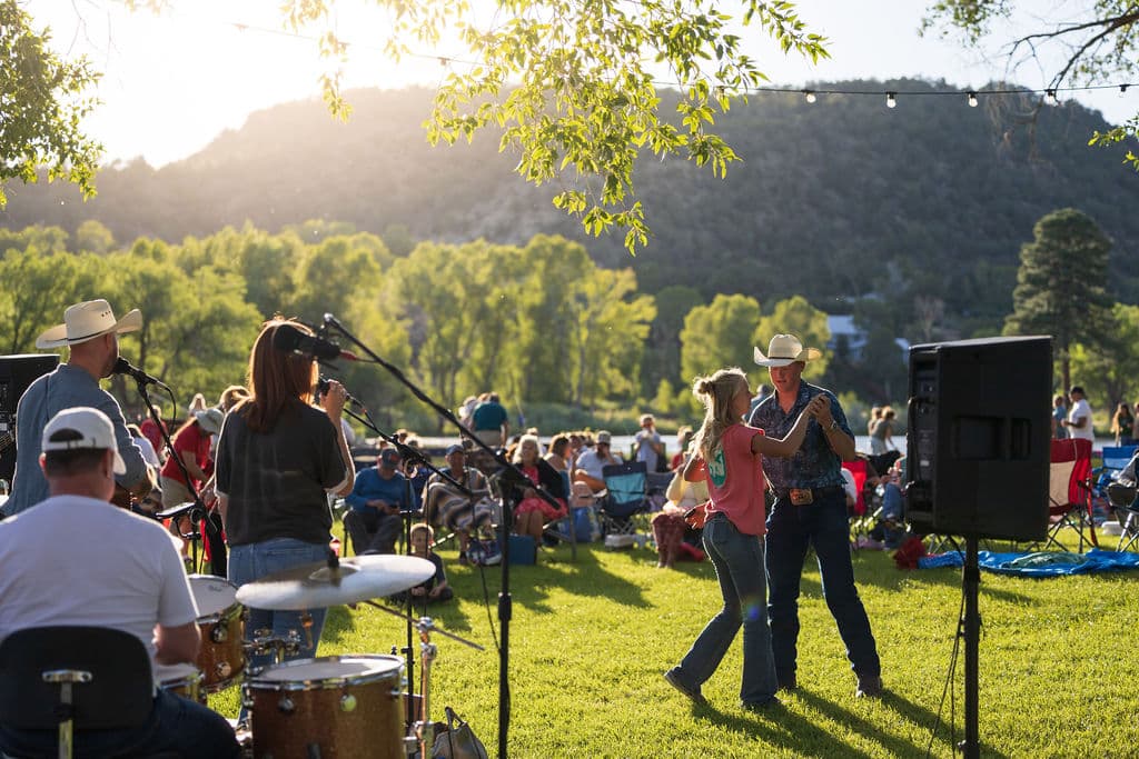 A couple in cowboy hats dancing on the grass in front of the band as the sun sets over the pond and hills