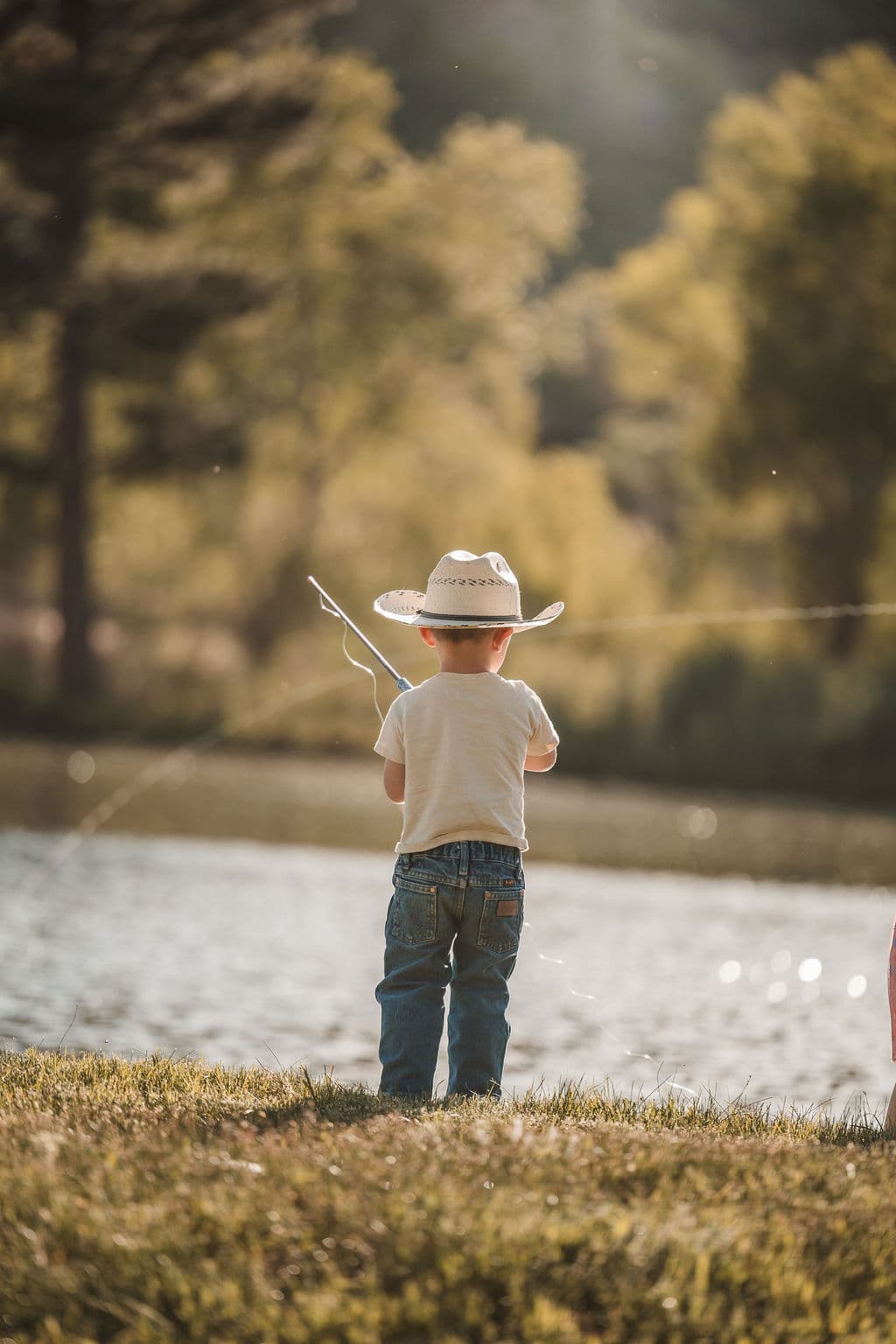 A young boy in a straw cowboy hat fishing at the water's edge, backlit by the warm evening sun
