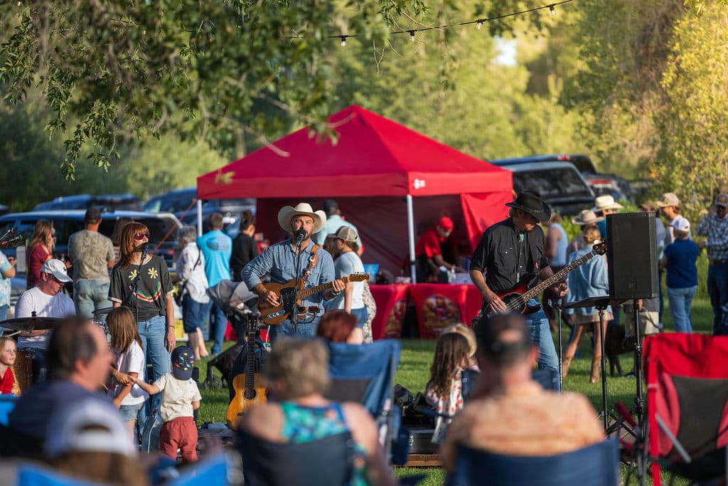 A cowboy-hatted singer and guitarist performing beside a red tent as families gather on the lawn beneath string lights at Locals Night