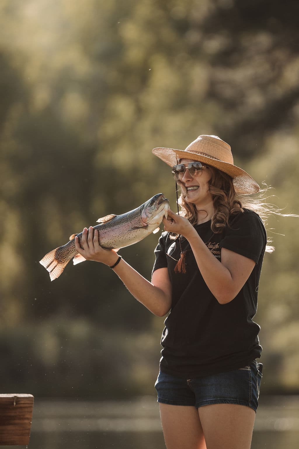 A smiling angler in a straw hat holding up a large rainbow trout caught at the pond