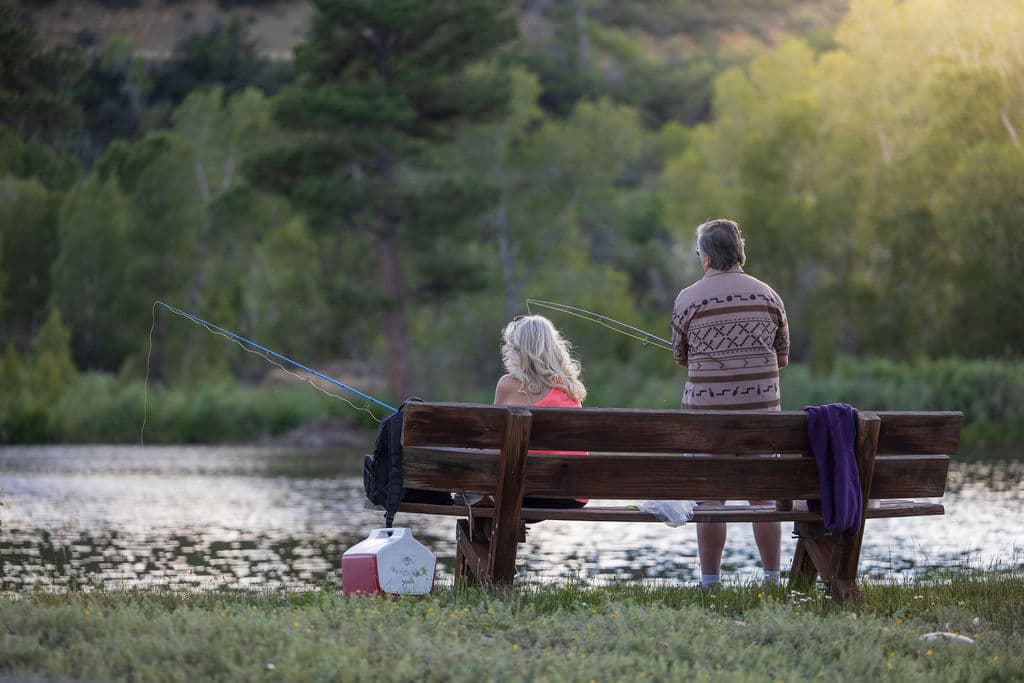 An older couple fishing together from a wooden bench at the pond's edge as the sun sets behind the trees