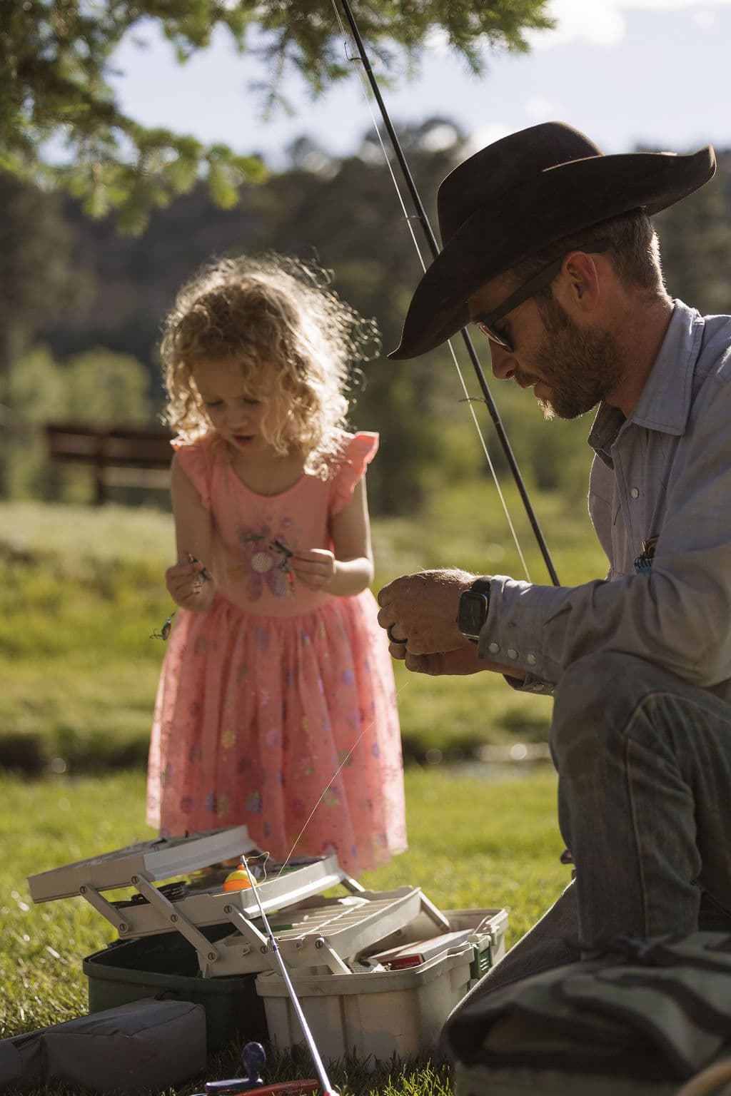 A cowboy kneeling to help a little girl in a pink dress rig her line at the tackle box before fishing
