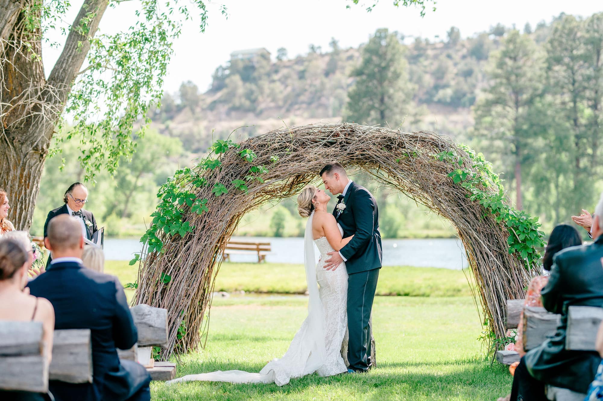 Bride and groom sharing their first kiss beneath the woven willow arbor with guests watching from rustic log benches