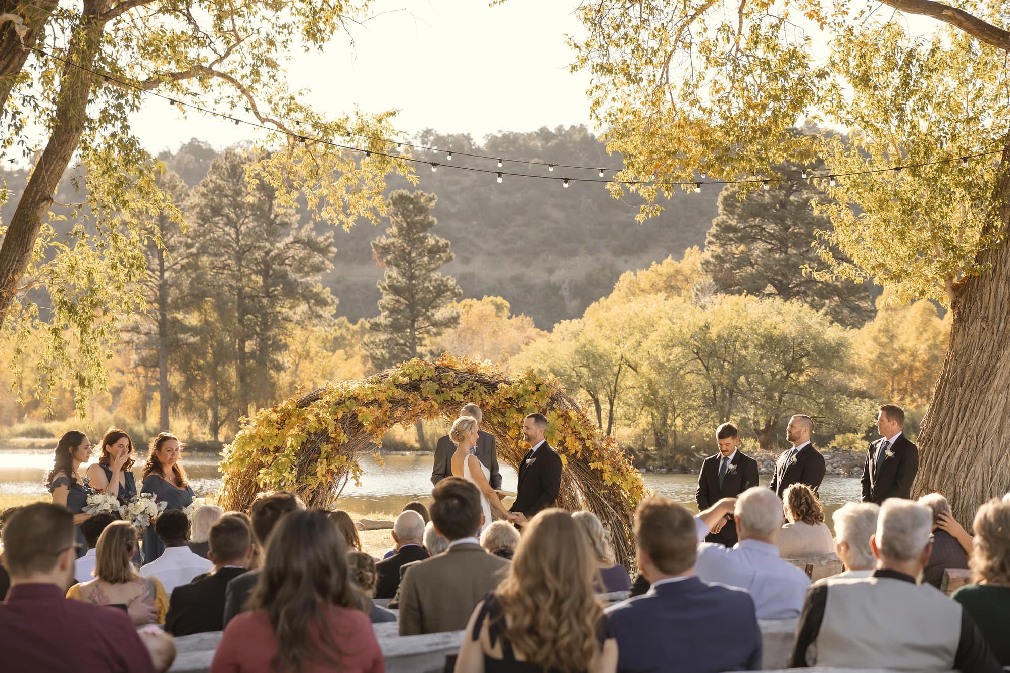 Autumn lakeside ceremony with grapevine arch, seated guests, golden foliage, and mountain backdrop
