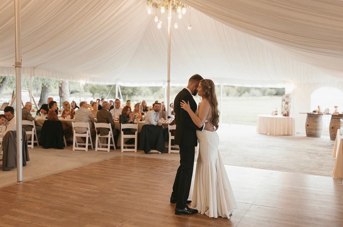 Couple sharing their first dance on the wood floor beneath draped ceiling and greenery chandeliers, guests and cake table visible in the background