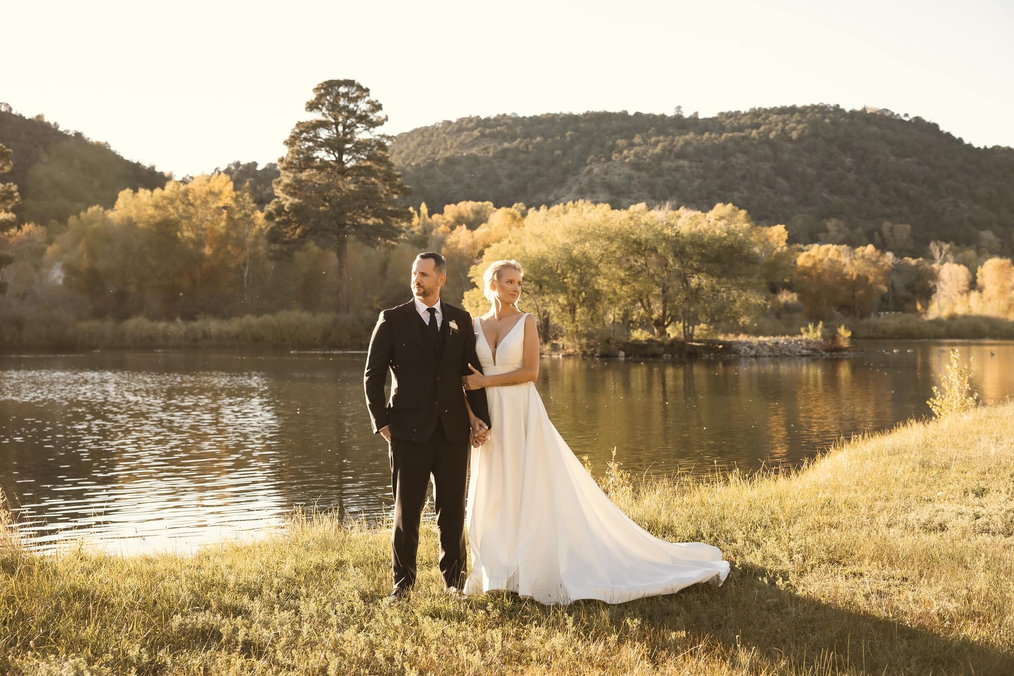 Megan and Eli standing together by the pond at golden hour with autumn foliage and mountain hillside behind