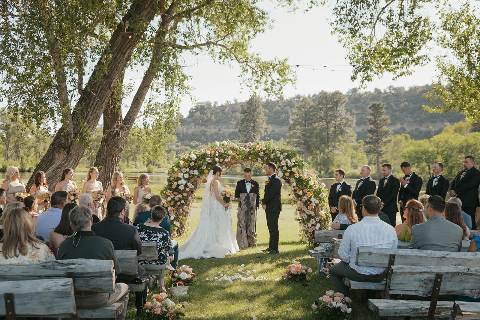 Bride and groom exchanging vows under a lush floral arbor beside the lake, with bridal party and guests seated on rustic log benches