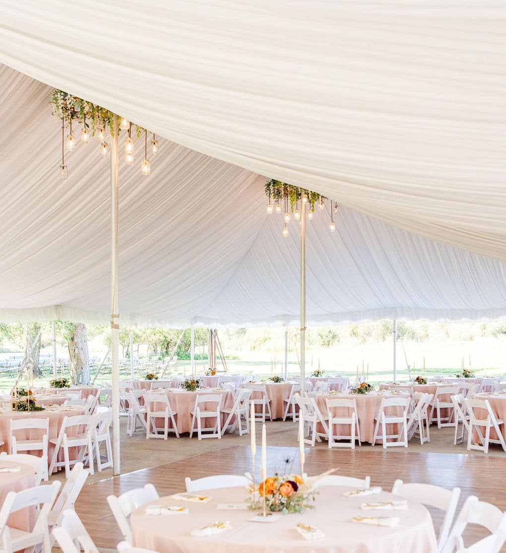 White draped reception tent interior with blush pink linens, white chairs, greenery chandeliers with Edison bulbs, dance floor, and views of the landscape