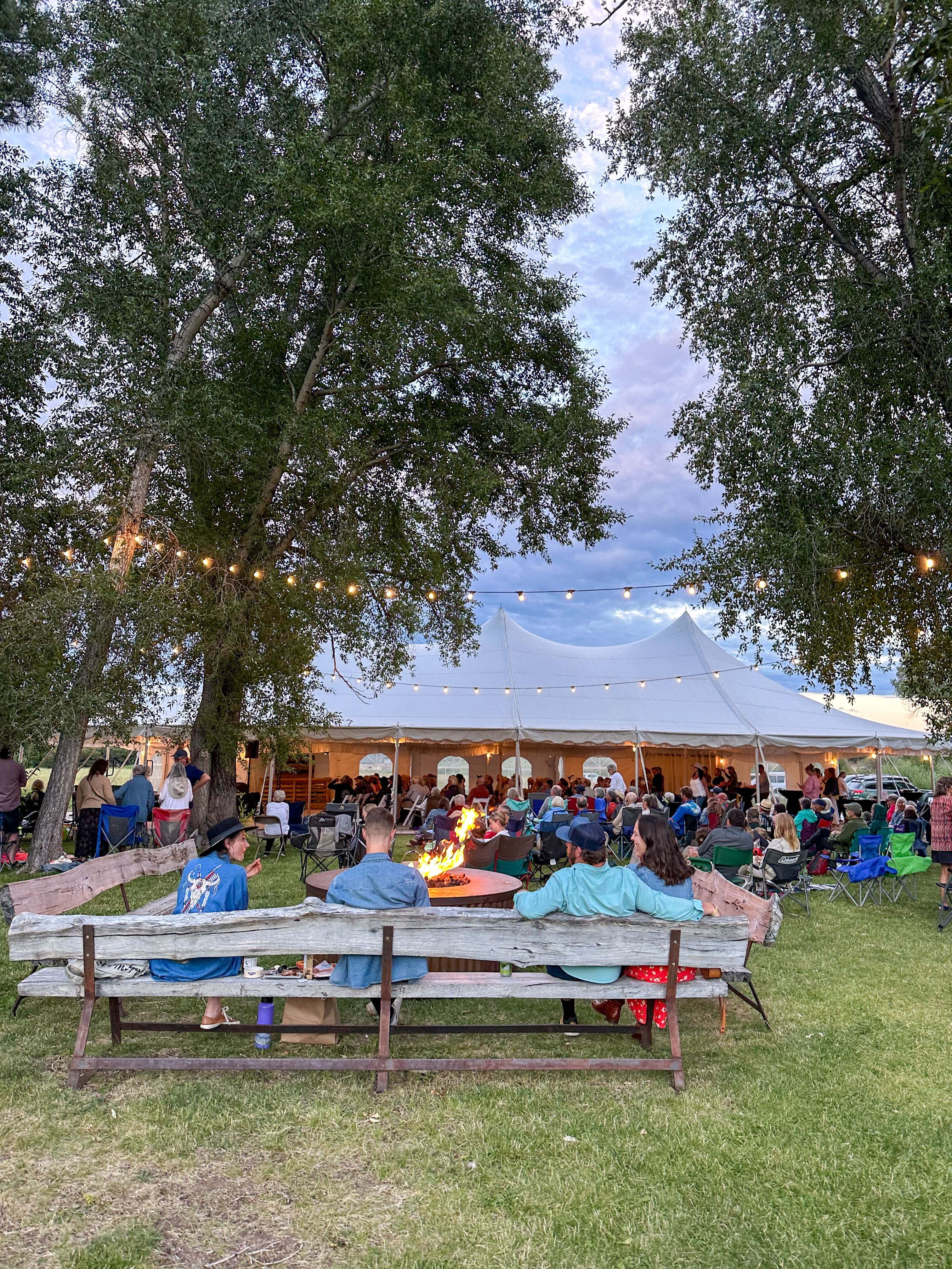 Community gathering around the firepit at dusk with string lights and the event tent glowing in the background