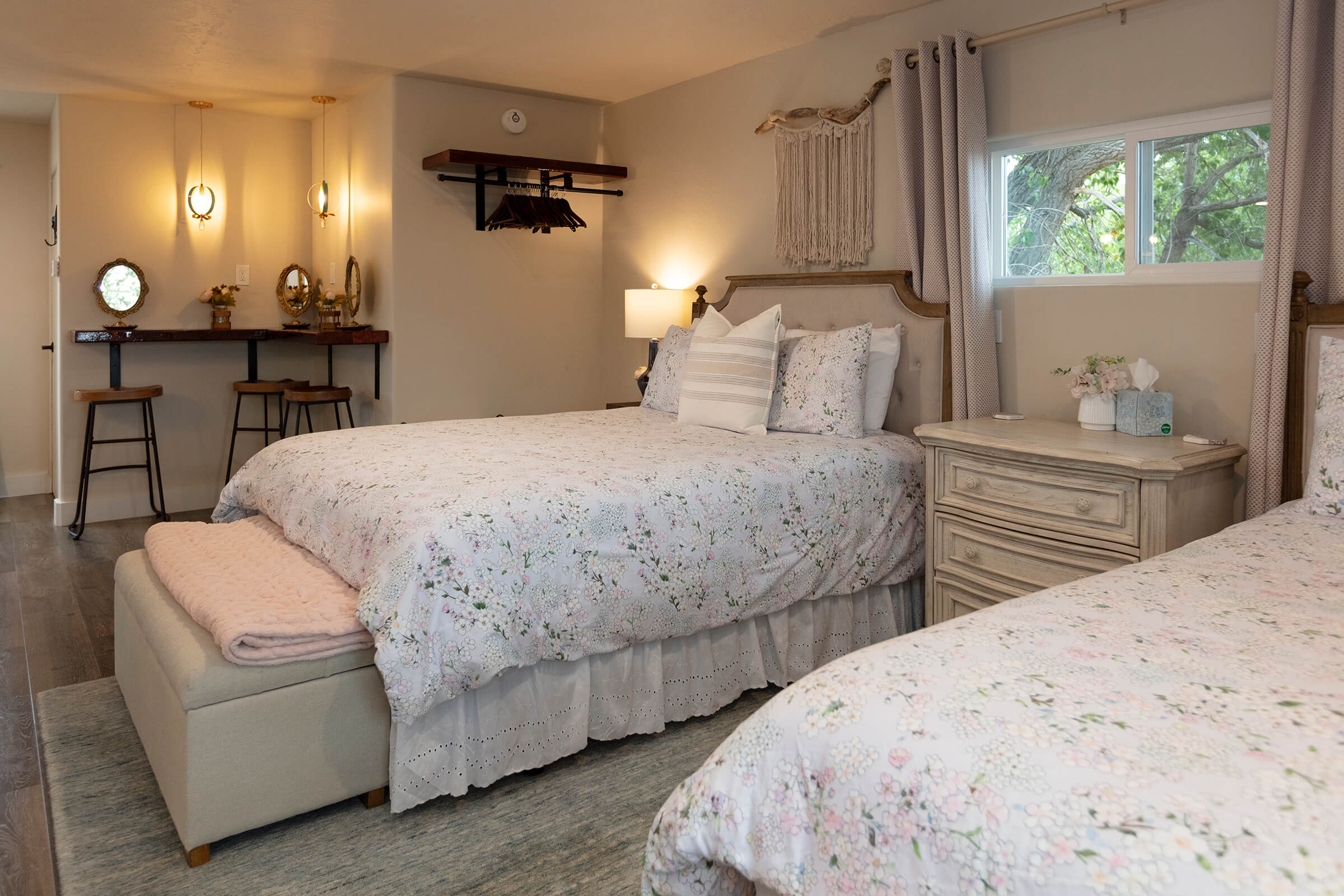 The Loft bedroom with two queen beds, floral bedding, vanity bar with illuminated mirrors, and macrame wall hanging