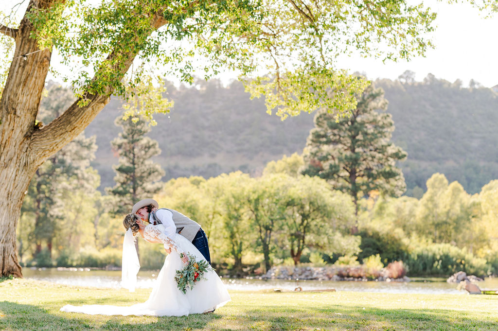 Couple sharing a romantic dip kiss on the lawn beside the lake with mountains and cottonwoods in the background