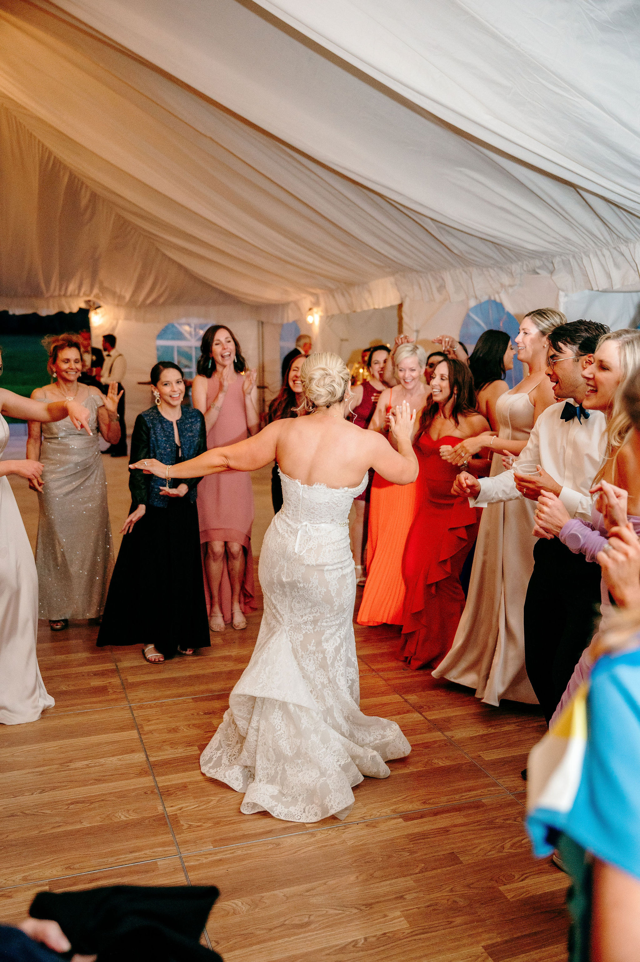 Bride dancing with arms outstretched surrounded by joyful guests on the wooden dance floor under the draped tent