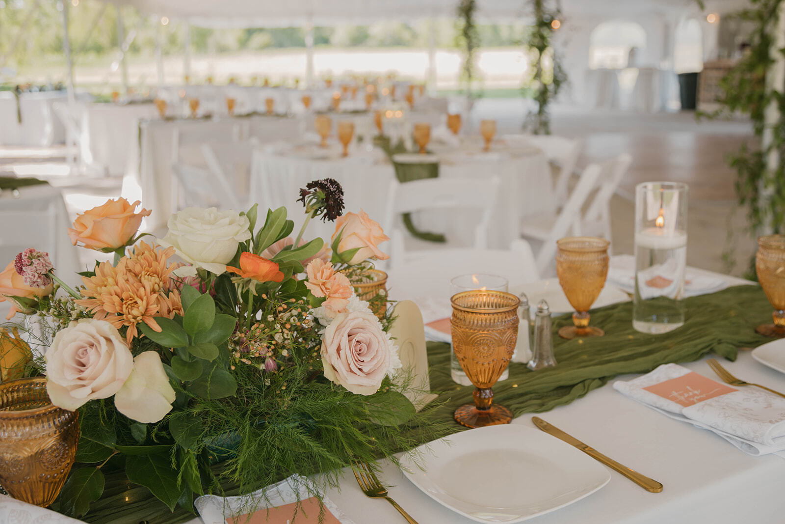 Refined reception tablescape with blush and peach roses, olive cheesecloth runner, amber goblets, and gold flatware