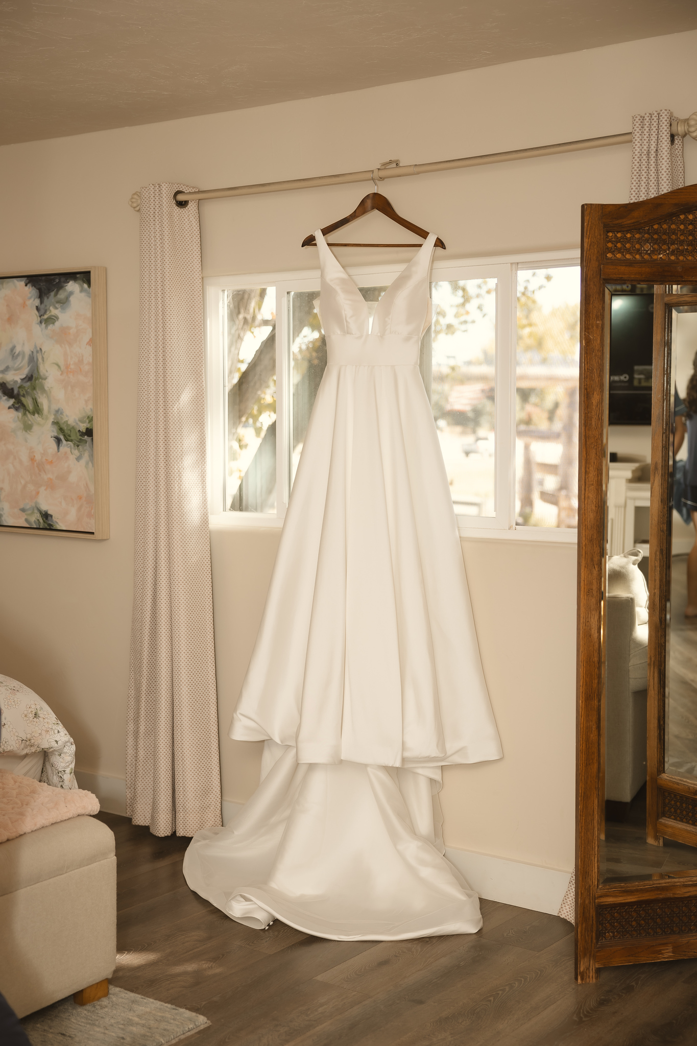 Bridal gown hanging in The Loft window with natural light, flanked by a floral painting and ornate standing mirror