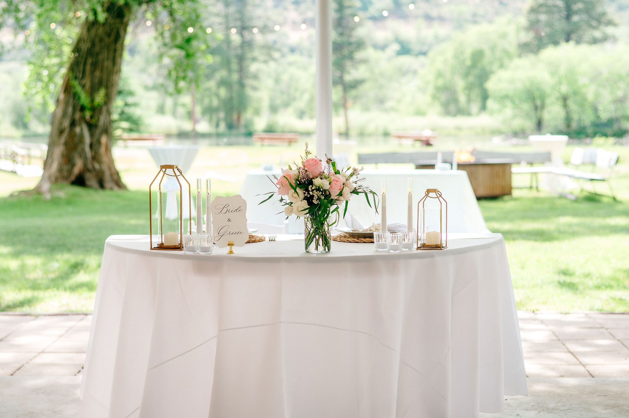 Sweetheart table styled with pink roses, gold lanterns, and taper candles with lake and fire pit visible in the background