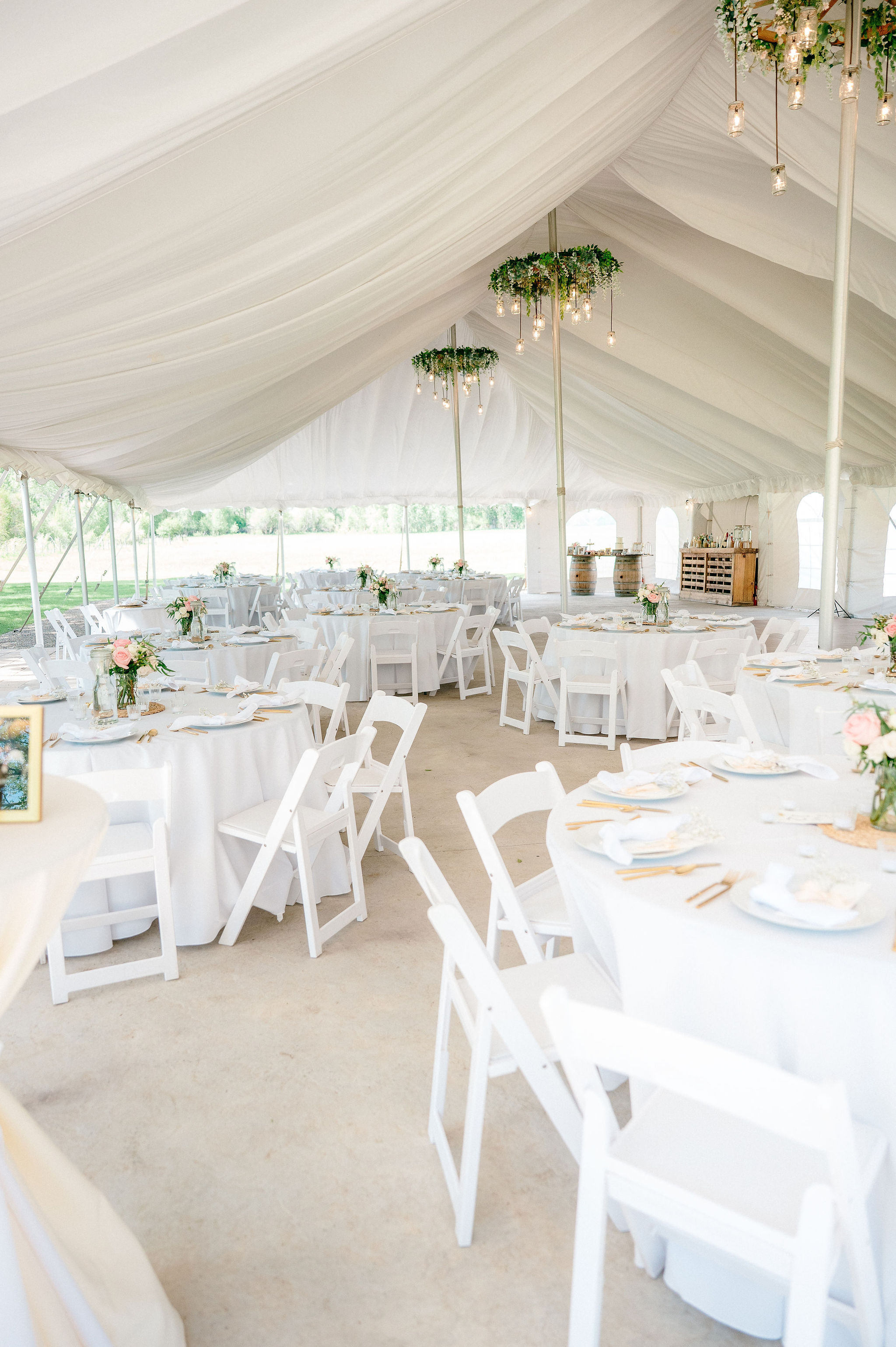 Full tent interior with white round tables, pink rose centerpieces, greenery chandeliers, barrel bar, and lake visible through open sides
