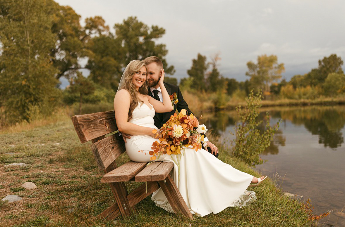 Katarina and Brandon sitting on a rustic bench by the pond at golden hour with an autumn dahlia bouquet