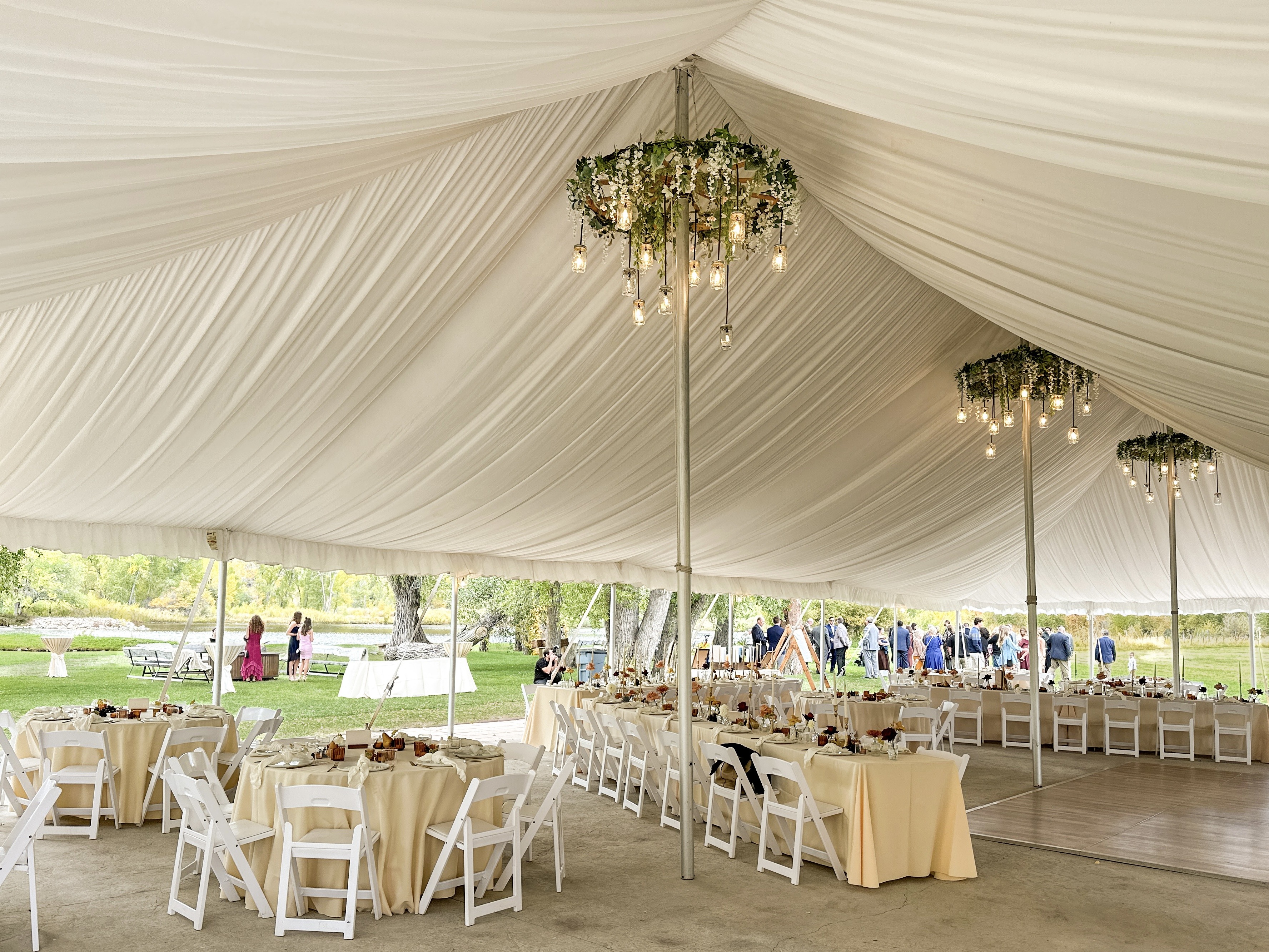 White draped reception tent interior with champagne linens, white chairs, greenery chandeliers with Edison bulbs, and guests mingling on the lawn